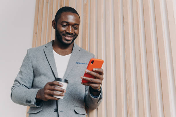 istock Enjoying coffee break at work. Young positive african man office worker in suit using mobile phone 1400396124