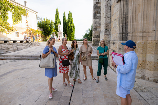 A wide angle side view of a group of women on a tour of a small historical village called Auvillar in Toulouse in the South of France. They are being shown round by a man who knows all about the history of the village.