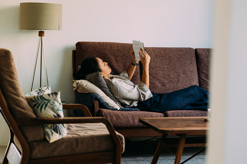 Young Asian woman enjoying her free time. She is in her living room, relaxing while reading a book.
