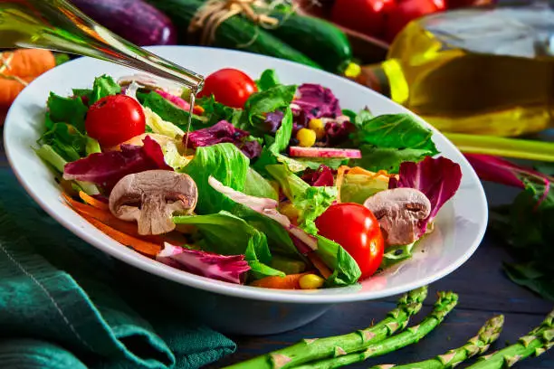 Vegan food themes. High angle view of a colorful spring salad on rustic white wood table. Included ingredients: Olive oil, peppermint, chia, tomatoes, broccoli, lettuce, bell peppers, mushroom, carrots, radicchio and almonds.