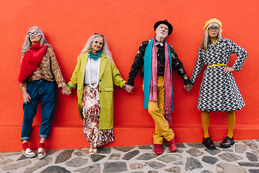 Senior friends holding hands while standing together against a red wall. Four elderly people expressing their unity and support of each other. Group of mature friends wearing colourful clothing.