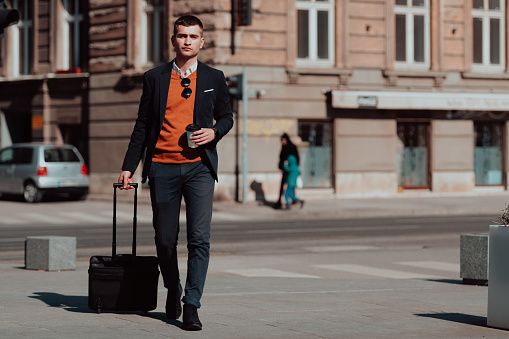 Going to the airport terminal. Young confident businessman traveler walking on city streets and pulling his suitcase drinking coffee and speaking on a smartphone. High quality photo