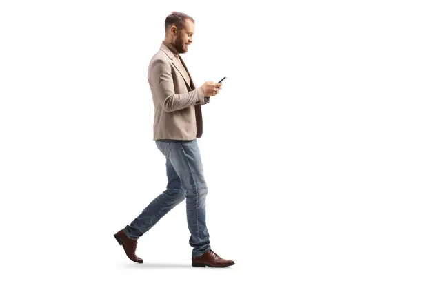 Full length profile shot of a man walking and looking at a smartphone isolated on white background