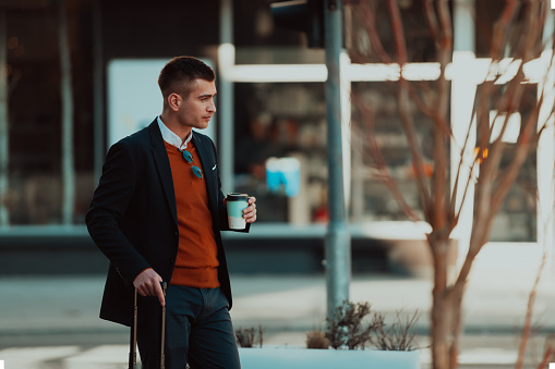 Going to the airport terminal. Young confident businessman traveler walking on city streets and pulling his suitcase drinking coffee and speaking on a smartphone. High quality photo