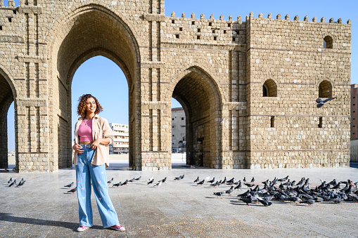 Full length portrait of casually dressed Middle Eastern woman and flock of pigeons in front of historical landmark at start of pilgrim’s road to Mecca.