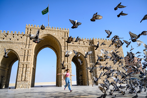 Full length view of playful woman running in front of 3-arched gateway landmark where Hajj pilgrims would begin their trek to Islam’s holiest city.