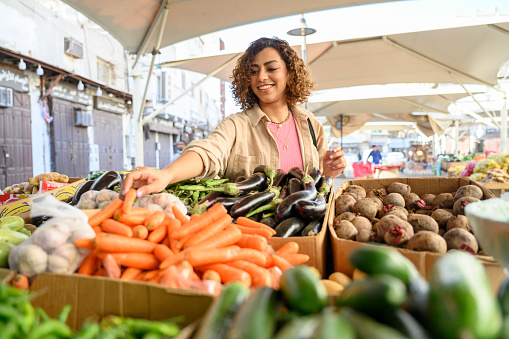 Middle Eastern woman in casual weekend attire selecting carrots from boxes of fresh vegetables under umbrella at open air food stall.