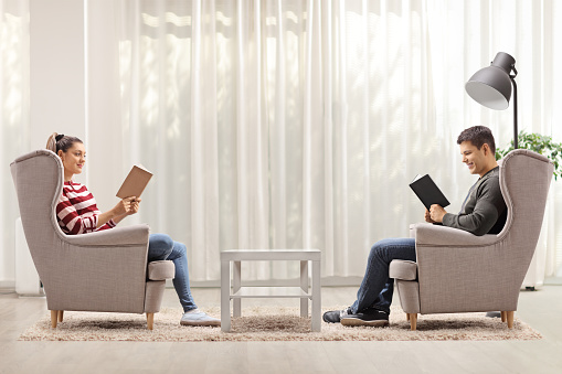 Profile shot of young man and woman sitting in armchairs and reading books at home