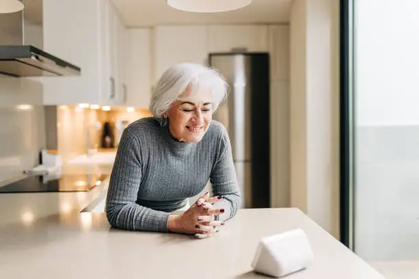 Smiling mature woman speaking to her home assistant on a smart speaker. Happy senior woman giving voice commands to her virtual assistant.