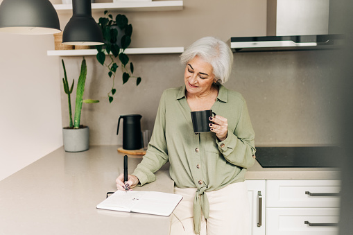 Carefree senior woman writing notes in her diary while standing in her kitchen at home. Happy mature woman making a to-do list while enjoying a cup of coffee.
