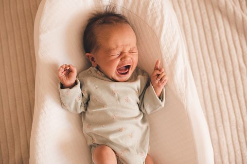 Unhappy sad baby lying in bed crib wake up and crying at home in room. Sad sick newborn child shouting with stomach pain and colics close up. Healthcare. Childhood.