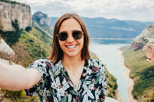 Portrait of young happy smiling woman with short brunette hair, floral shirt and sunglasses, taking selfie with smartphone, over the top of the mountain lake. Adventure, travel, holiday concept.