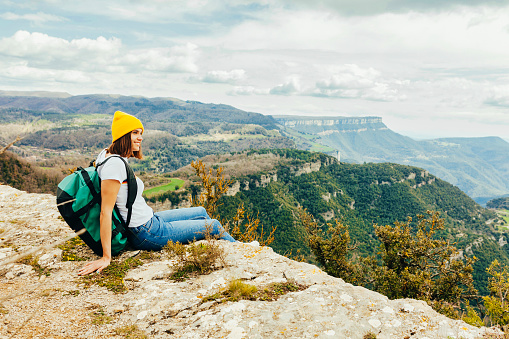 Young happy smiling woman with a backpack and yellow beanie, sitting on the edge of a rock and looking at the sky with clouds. Adventure, travel, holiday concept.