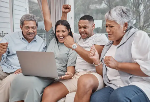 Photo of Happy and affectionate mixed race family of four using a laptop to watch sports and cheer on their favourite team in the home living room. Married couple sitting with their mature parents on the sofa