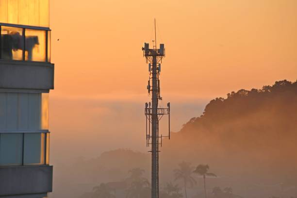 Tall telecommunication tower in the city, next to a building. Scenery with fog, diffuse, of poor visibility, with ray of the rising sun. In Background silhouette of mountains.