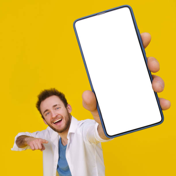 istock Young happy man holding smartphone showing a white empty screen check this out, cellphone display mock up mobile app advertisement. isolated on yellow background, Product placement 1400423196
