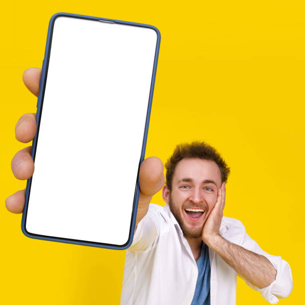 istock Great offer. Young happy man holding smartphone showing a white empty screen and exciting to win isolated over yellow background, celebrating success. Product placement for mobile app advertisement 1400423197
