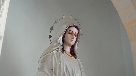 Statue of the Virgin Mary standing indoors with her head bowed looks straight