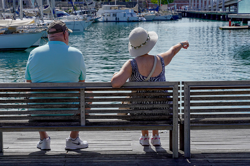 Barcelona, Spain - May 26, 2022: Older couple contemplate sitting on a bench next to the marina, the woman points with her finger at a point of interest that they both look at, they are wearing comfortable sports shoes.