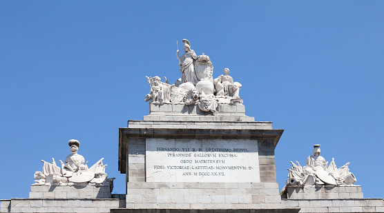 The top of the Toledo Gate, Puerto de Toledo, in Madrid, Spain, which was completed in 1827. The attic has a dedication to King Fernando VII. The central sculpture represents the power of the Spanish monarchy.