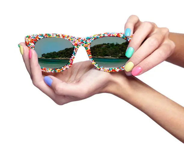 Woman is holding fancy sunglasses with sea beach reflection. Female fingers with bright green, yellow, pink and blue nails manicure. Girl's hands with glasses isolated on white background