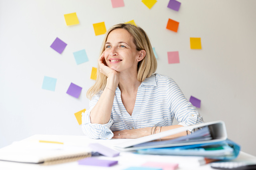 Portrait of pretty young blonde woman with beautiful laugh sitting at office table in front of white wall with colorful sticky notes