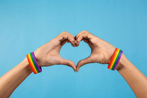 Hand making heart sign with gay pride LGBT and rainbow-patterned wristband on wrists, on blue color background. LGBT and love concept