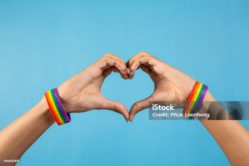 Hand making heart sign with gay pride LGBT and rainbow-patterned wristband on wrists, on blue color background. LGBT and love concept Rainbow Flag Stock Photo