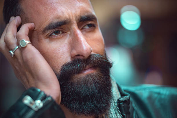 In this headshot with copy space, an Asian/Indian tensed and depressed young man with a long beard and mustache looks away, contemplates, and holds his head.