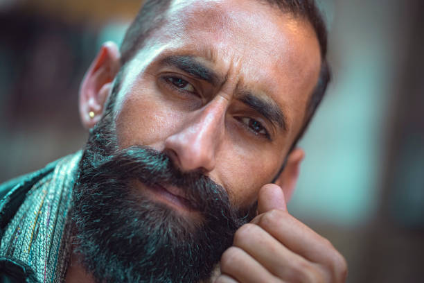 In this headshot image, an Asian/Indian tensed and depressed young man with a long beard and mustache looks at the camera.