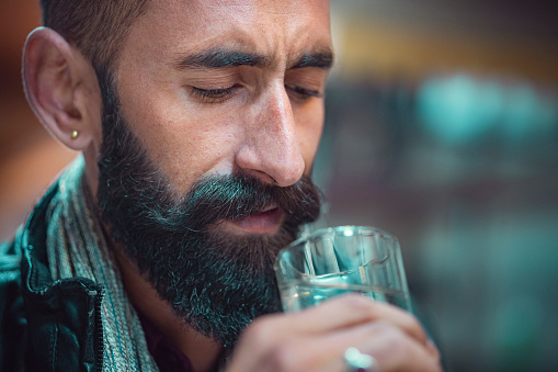 In this headshot with copy space, an Asian/Indian young man with a long beard and mustache drinks a glass of water as he feels tensed and exhausted.