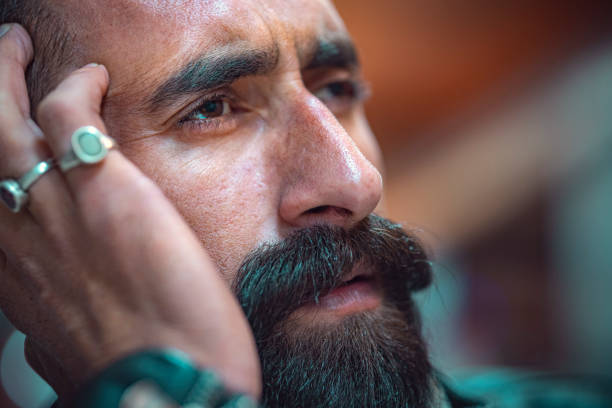 In this headshot with copy space, an Asian/Indian tensed and depressed young man with a long beard and mustache looks away, contemplates, and holds his head.