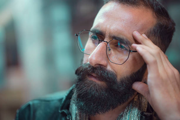 In this headshot with copy space, an Asian/Indian young man with a long beard and mustache contemplates and looks away.