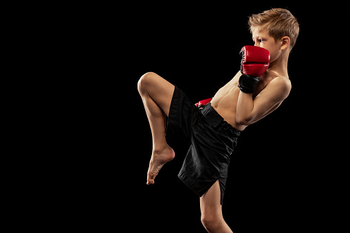 Studio shot of preschool boy, cute kid in sports shots and gloves training alone on black background. Beginner fighter of mma in action, motion, sport, challenges concept. Copy space for ad