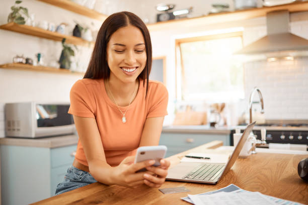 istock Young beautiful happy mixed race businesswoman holding and using a phone while working from home. One content hispanic woman using social media on her cellphone while using a laptop sitting at a kitchen counter 1400435777