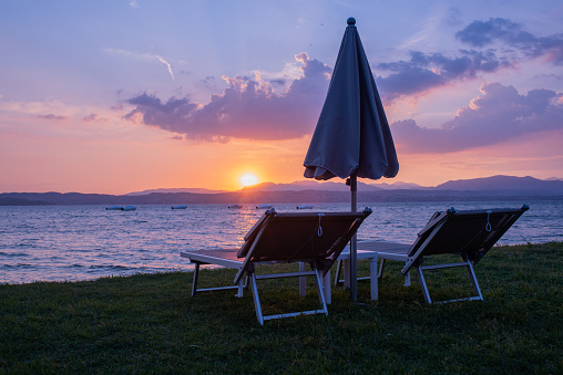 Deck chairs with umbrella on lake garda are waiting for tourists. Moody sunset. Background for vacation and relaxation.