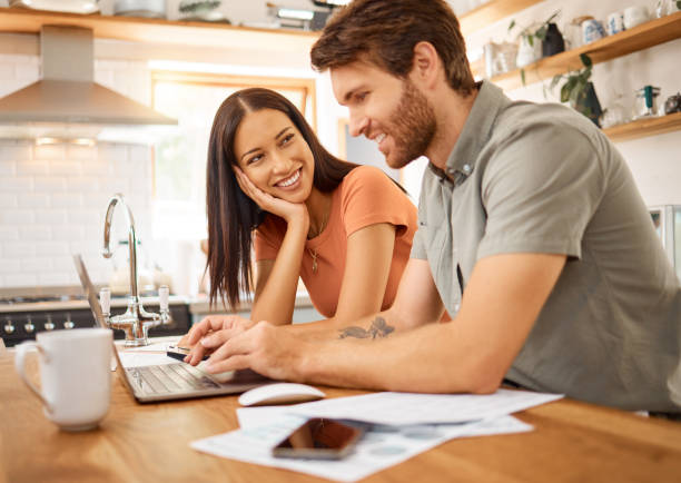 istock Young cheerful interracial couple bonding while working on a laptop together at home. Caucasian boyfriend and girlfriend working through their bills and using a laptop. Content wife staring at her husband while typing on a computer 1400436736