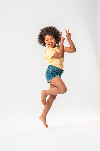 Black-haired little girl of African origins wearing jeans shorts and a yellow shirt, jumping high without shoes in front of a white studio background. Signaling peace signs with both her hands and slightly sticking the tongue out. Looking at camera.