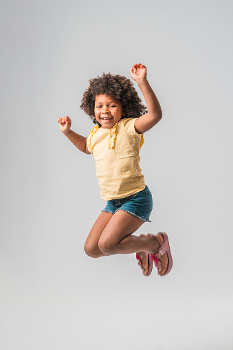 Little Caribbean girl in summer wear, jumping high  with legs bent in the knees and pulled up, arms lifted up, wearing pink sandals. Looking at the camera with a toothy smile and her hair flying up.