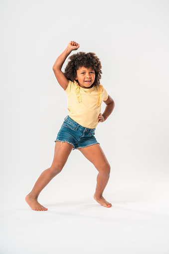 Small African girl wearing jeans shorts and a yellow t-shirt, dancing barefoot in front of a white studio background. One arm up in the air, the other one on the hip, legs doing the twist moves. Looking at camera.