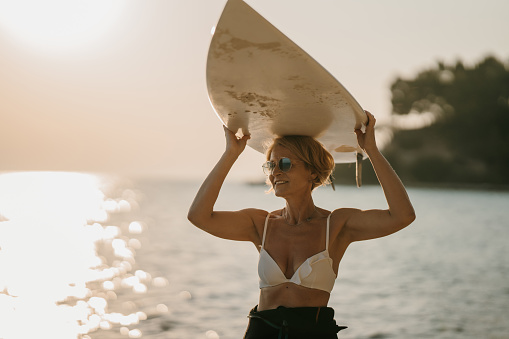 Mature woman surfer standing on the beach by the sea and holding her surfboard over her head.