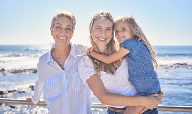 portrait of female family members posing together at the beach on a sunny day. grandmother, mother and granddaughter standing together on seaside promenade. multi-generation family of women and little girl spending time together - blond hair carrying little girls small imagens e fotografias de stock