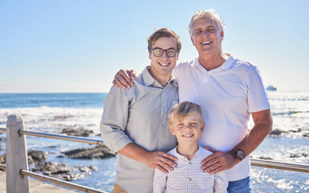 male family members posing together at the beach on a sunny day. grandfather, father and grandson standing together on seaside promenade. multi-generation family of men and little boy spending time together - child little boys male caucasian imagens e fotografias de stock