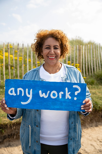 A senior woman seeking work, holding up a sign with 'any work?' written on it while looking at the camera and smiling. She is standing near Beadnell beach, North East England with some of the beach and a grassy bank behind her.