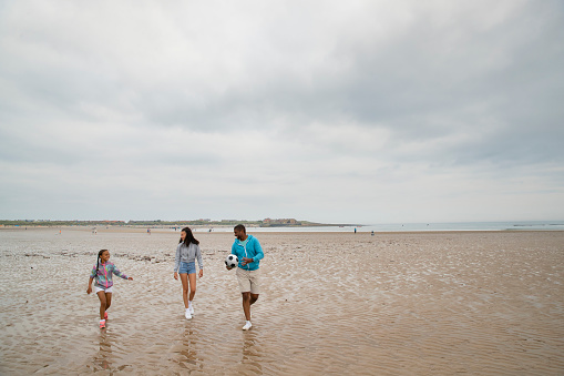 Two girls and their father walking together on Beadnell beach, North East England. The man is carrying a football and they are getting ready to have a game.