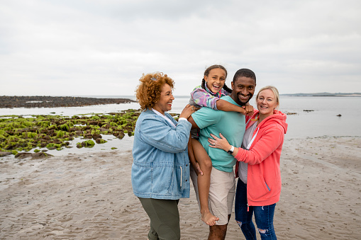 A multi-generation family standing together on Beadnell beach, North East England while looking at the camera and smiling. The man is giving his daughter a piggyback and the grandmother is tickling the young girl.