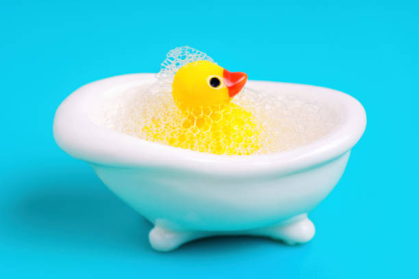 Toy yellow duck in a miniature white bathtub covered with soap bubbles. Bath time concept.