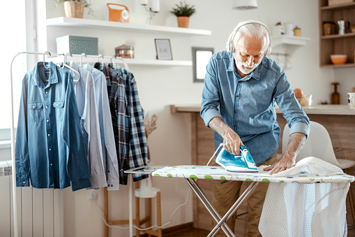 Bearded senior man listening to music while ironing at home