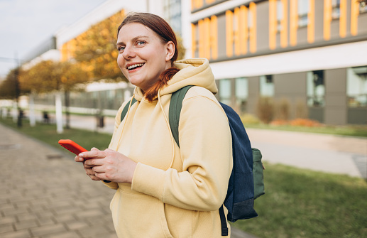 A portrait of a smiling beautiful woman texting sms with her phone on urban background. Happy student with backpack is using a smartphone outdoors, spring or autumn time.