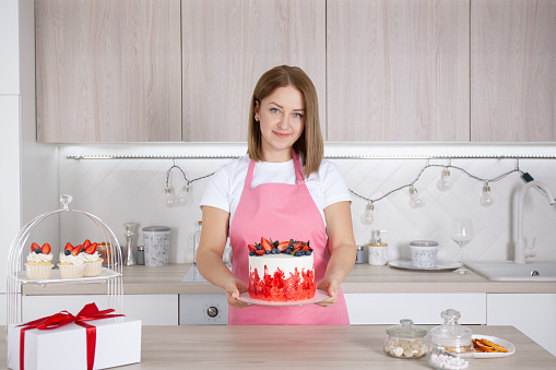 Pastry chef confectioner young caucasian woman in the kitchen. She holds out the cake on a plate. Nearby are cakes, a cake box and sugar in a sugar bowl